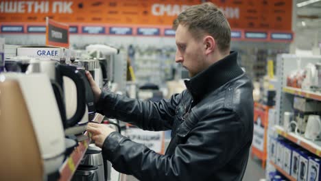 Young-man-in-hypermarket-choosing-good-electric-kettle-for-his-new-house-in-a-big-shop-with-is-a-big-choice-of-small-household-appliances