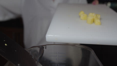 Amazing-slow-motion-shot-of-a-group-of-small-pieces-of-cut-pineapple-on-a-white-chopping-board-being-pushed-with-a-knife-into-a-blender-to-prepare-a-piña-colada-cocktail