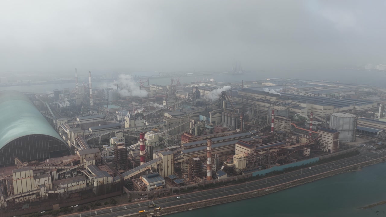 Expansive industrial plant with tall chimneys emitting white smoke and steam, contributing to air pollution and global warming under a gray, smoggy sky next to a river
