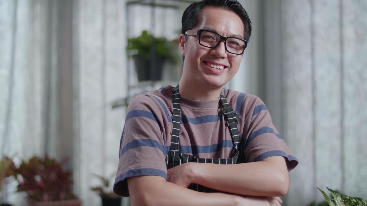 식물이 있는 아시아인 남자가 팔을 고 집에서 카메라에 미소 짓고 있습니다.