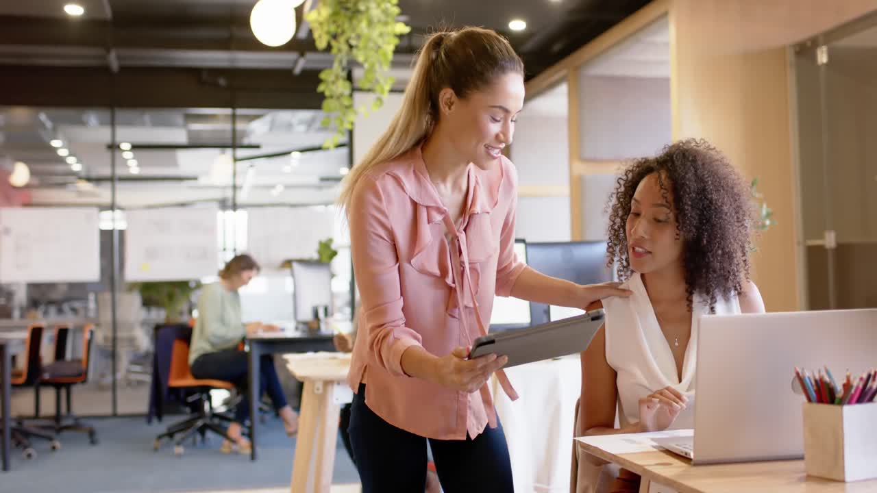 Happy diverse businesswomen discussing work and using tablet at desk in office, in slow motion