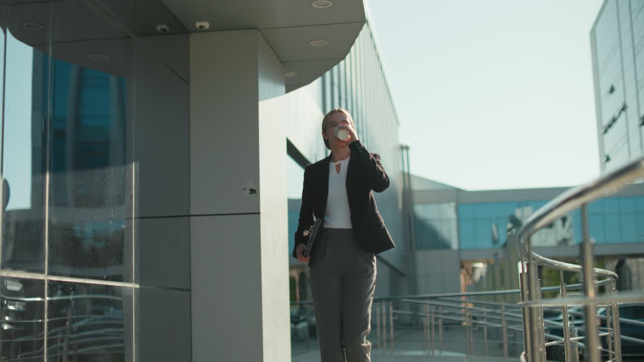 Young woman walking in heels holding tablet in right hand and coffee in left hand as she sips while passing modern building with parked cars in surrounding
