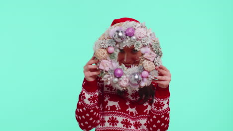 Adult-smiling-girl-in-red-Christmas-holiday-sweater-hold-raise-point-finger-on-festive-wreath-toy