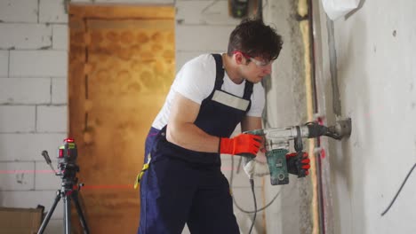 electrician man boring hole in wall for socket using laser level, working power drill, jackhammer with cylindrical nozzle. preparation for instsling electrical wires, construction work in apartment