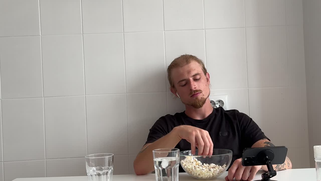 Молодой человек ест попкорн и смотрит телефон дома.