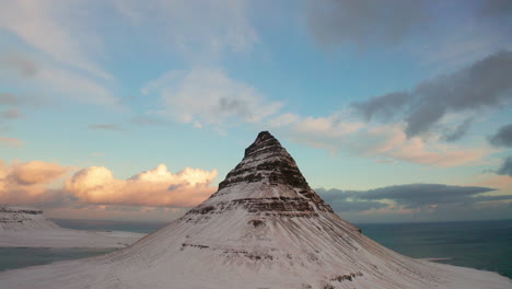 kirkjufell mountain iceland in winter, covered with snow sharpened peak at the top with long high curved sides, aerial panoramic descending view, blue cloudy sky at sunset in background