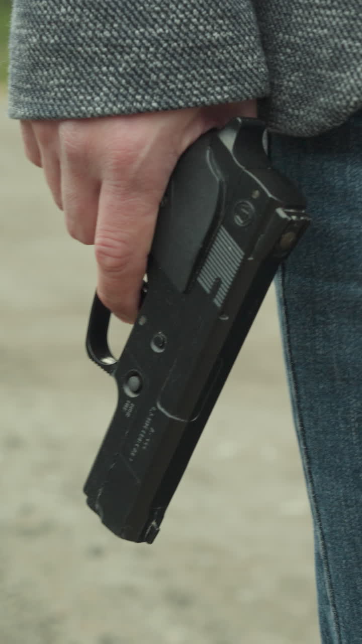 una vista de cerca de un hombre en vaqueros azules y una chaqueta gris sosteniendo una pistola negra a su lado mientras camina por un camino de tierra