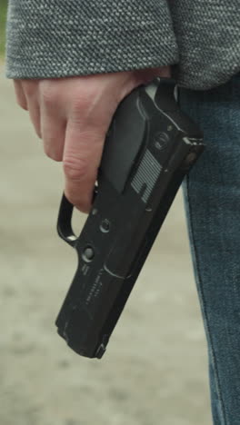 una vista de cerca de un hombre en vaqueros azules y una chaqueta gris sosteniendo una pistola negra a su lado mientras camina por un camino de tierra