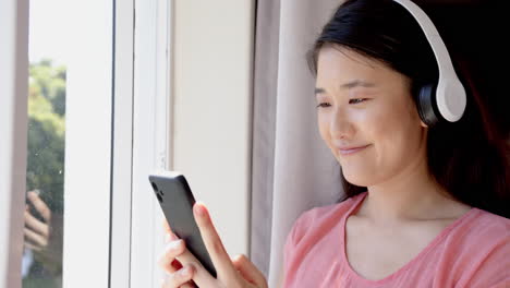 Listening to music, woman using smartphone and wearing headphones by window