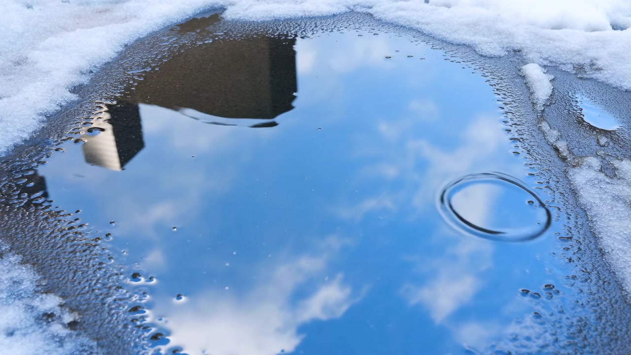 Gjennom denne sekvensen forblir en grunn sølepytt sentrert i rammen, mens blå himmel og myke skyer reflekteres på overflaten, med smeltende snø
