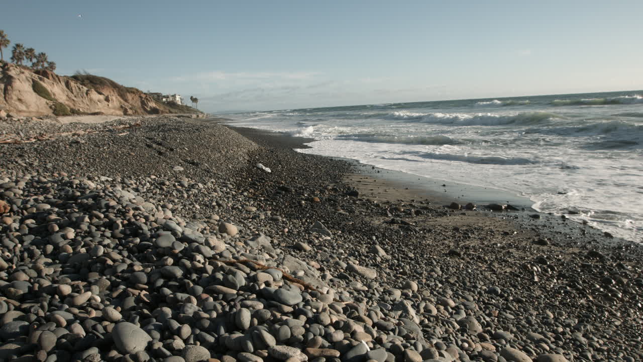 Gentle ocean waves roll onto a tranquil shore, framed by scattered pebbles on the sand. Soft light and rhythmic surf create a calming, meditative scene perfect for nature and relaxation themes.