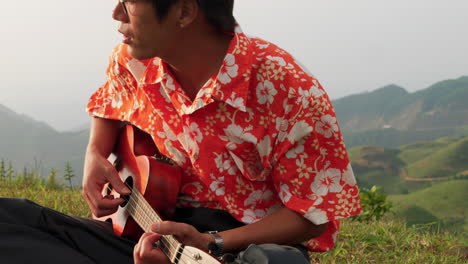 close up shot of an asia man sitting on the top on a mountain and playing ukulele while on a trip with mountain range in the background