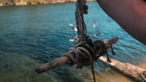 close up: hand holds the end of the rope swing with wooden sticks on the river bank