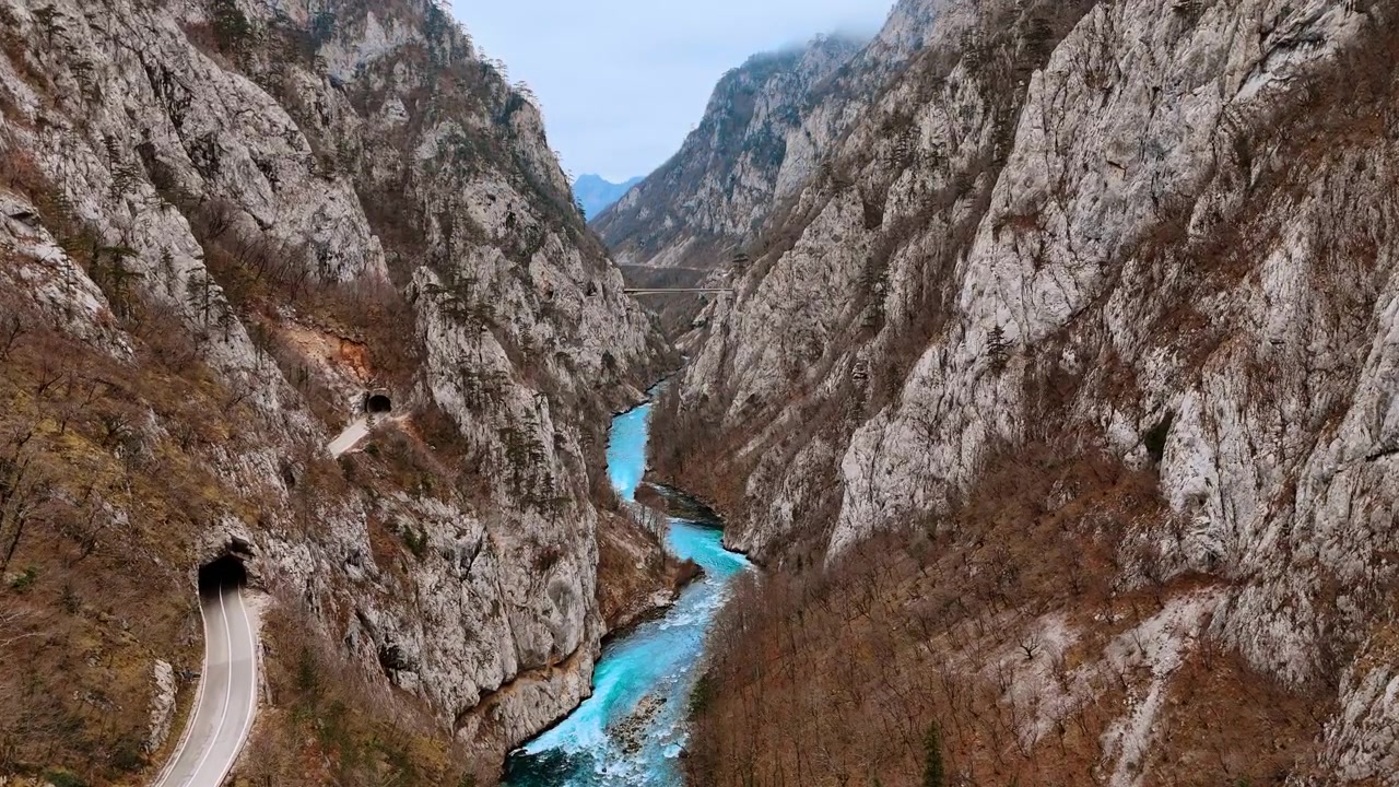 Vista aerea di una strada nel canyon con fiume turchese e tunnel e un ponte distante. Una strada entra in un tunnel roccioso sulla scogliera sinistra mentre il fiume turchese scorre sotto e un ponte attraversa la gola più avanti.