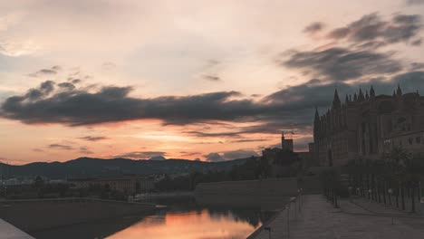 golden sunset over palma cathedral with dramatic clouds and mountains (palma, mallorca, balearic islands, spain). sunset transitions to night with tranquil reflections in the water (timelapse)