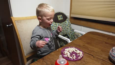 Toma-Amplia-De-Mano-De-Un-Niño-Comiendo-Arroz-Y-Carne-Con-Una-Cuchara-De-Metal-Sentado-En-La-Mesa-De-La-Cocina