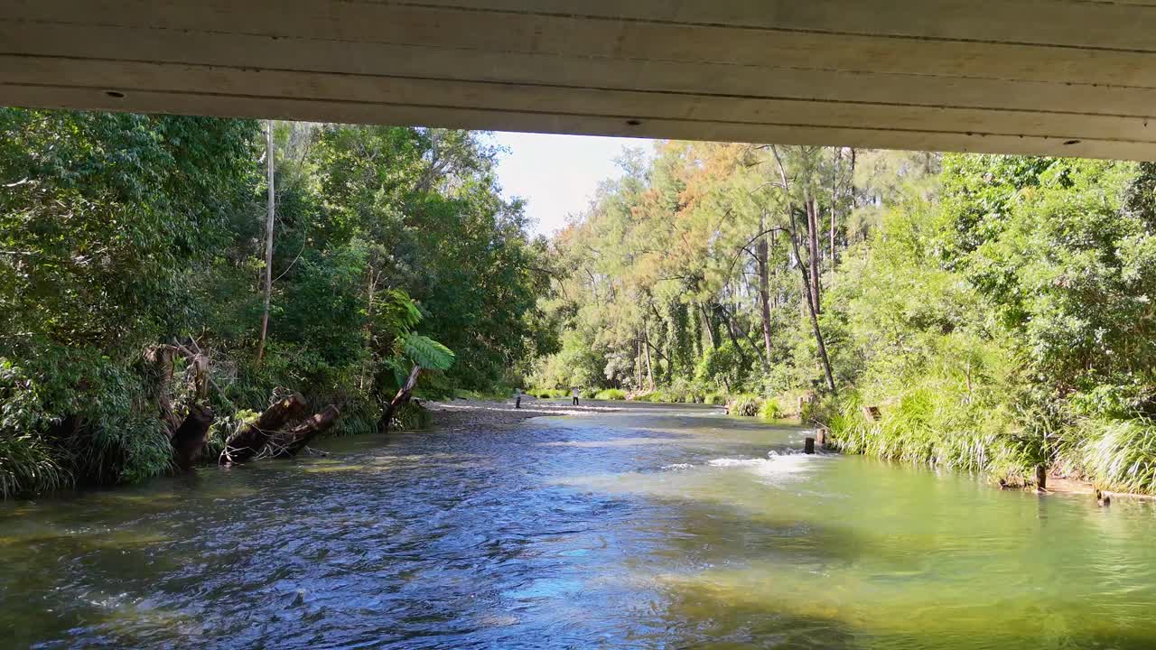 A tranquil river flows under a bridge in lush Bellingen, NSW. Sunlight filters through trees, creating a peaceful, natural scene