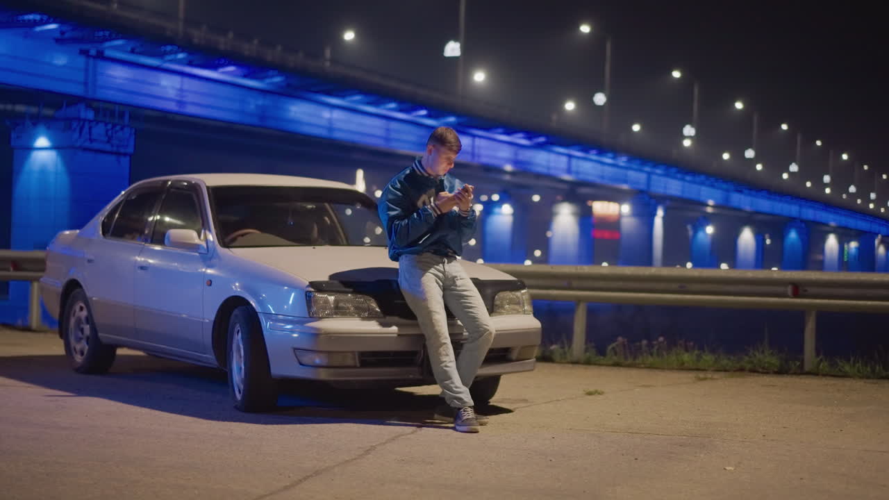 hombre blanco apoyado en un coche bajo un puente iluminado de noche realizando una reparación en la carretera, chispas del motor, luces azules reflectantes, chaqueta vaquera, smartphone cerca, ambiente cinematográfico y solitario