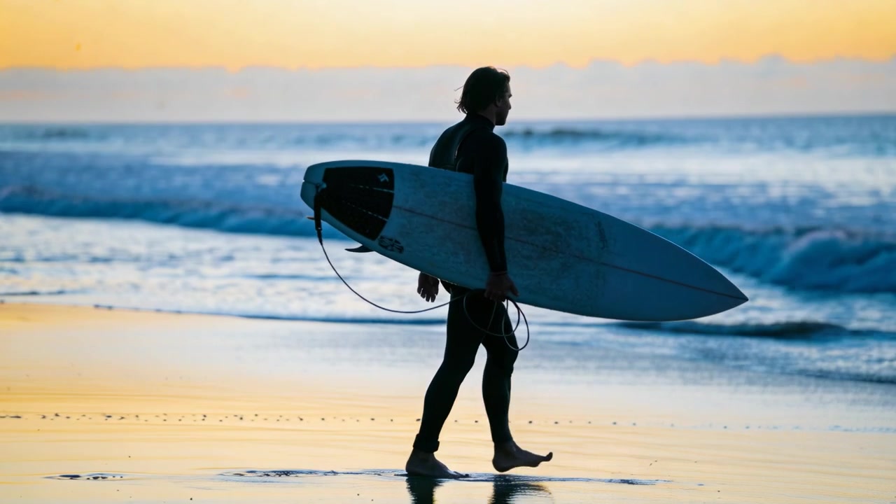 Walking male surfer in black wetsuit carrying surfboard along shoreline, breaking waves drawing him