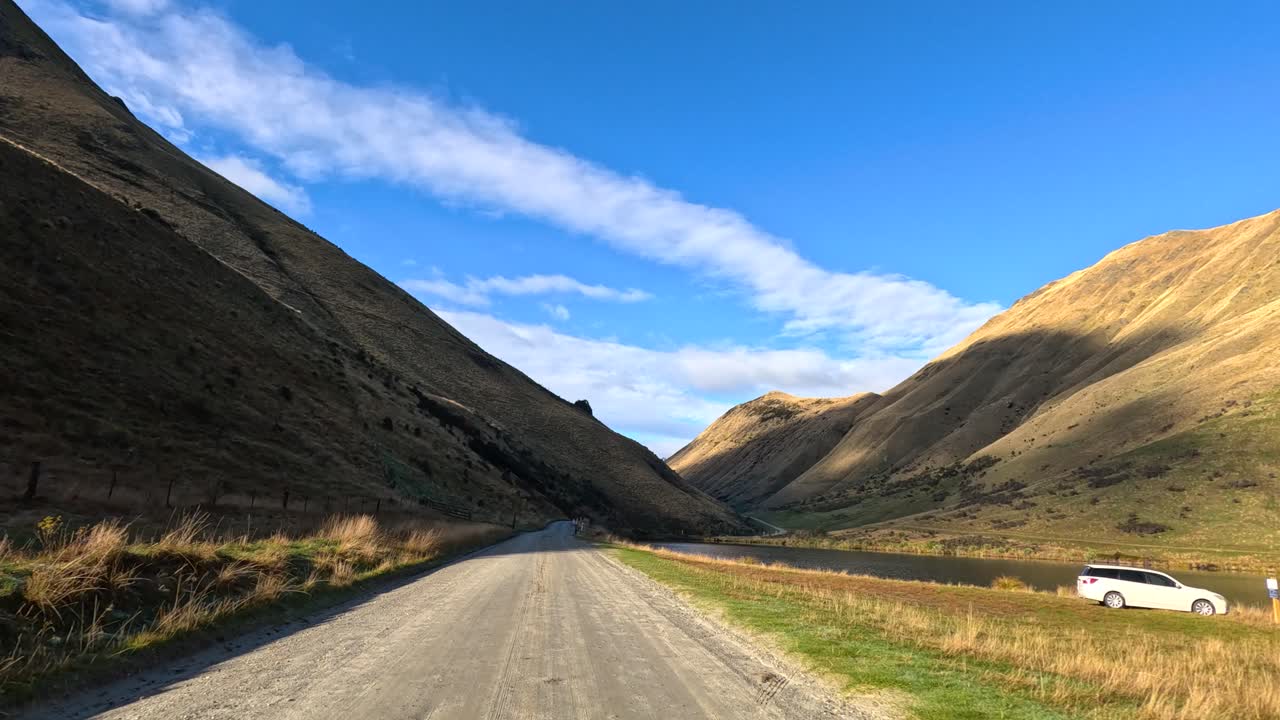 Vehicle drives on gravel road between sunlit hills under clear blue sky, camera moves forward