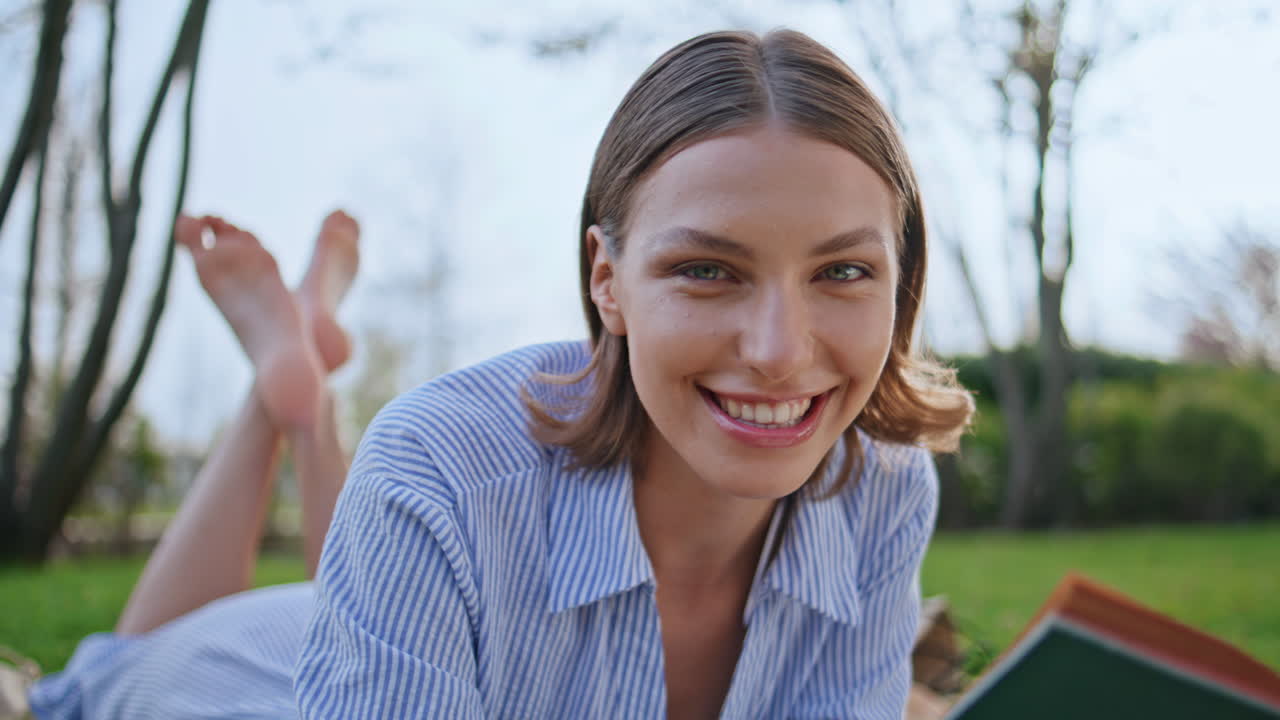 Joyful girl holding novel lying on picnic blanket at meadow closeup