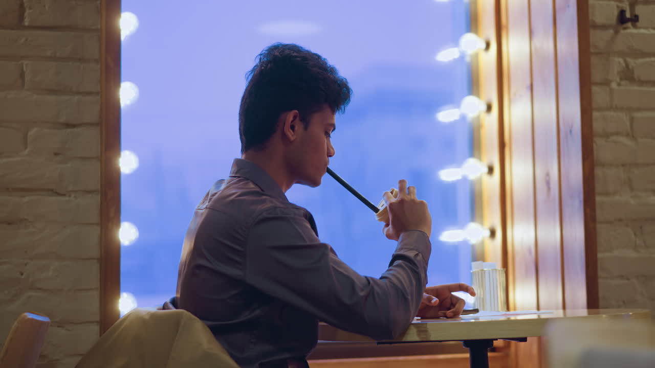 Young man wearing gray shirt sitting by window with soft glowing lights, sipping from disposable cup through long straw, enjoying drink in quiet cozy cafe during evening with warm urban ambiance