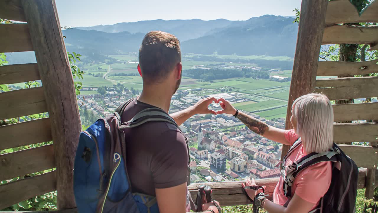 Young hiker couple forms common heart with their fingers at viewpoint over village in valley. Making Heart sign with hands at panoramic viewing point