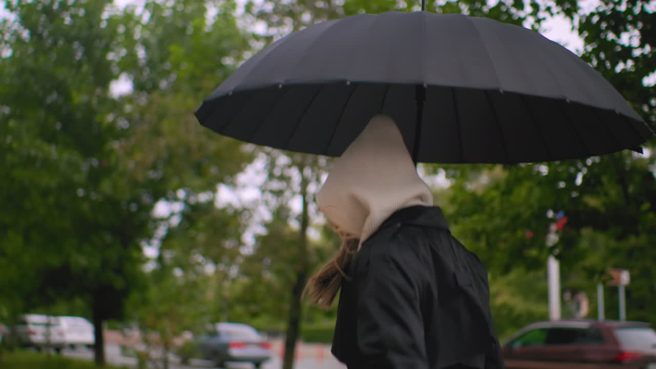 Back shot of woman in long black trench coat running along city sidewalk on windy autumn day, belt tie swinging freely, blurred cars and trees in background creating urban seasonal atmosphere