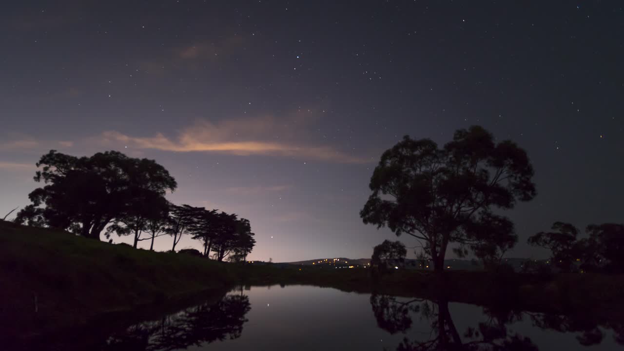 전경에 고무 나무가 있고 댐 물에 밤하늘이 반사되는 호주 시골 풍경 위에 별들의 시간 경과