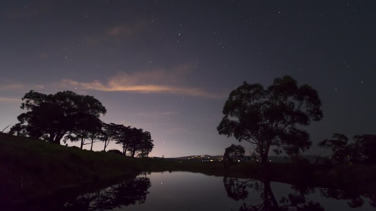 A time lapse of the stars over rural Australian landscape with a gum trees in the foreground and the reflection of the night sky in the water of a dam