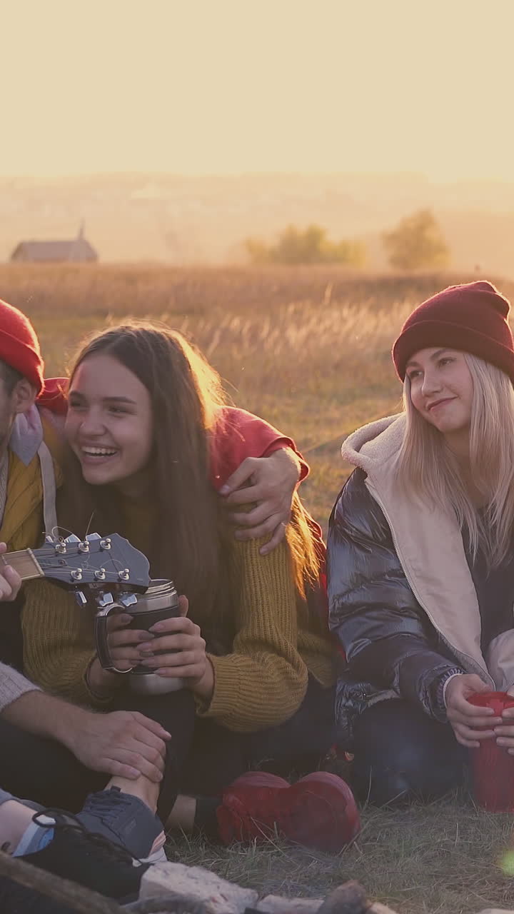 group of happy friends rests listening to guitar playing at bonfire in camp with tent in warm autumn evening
