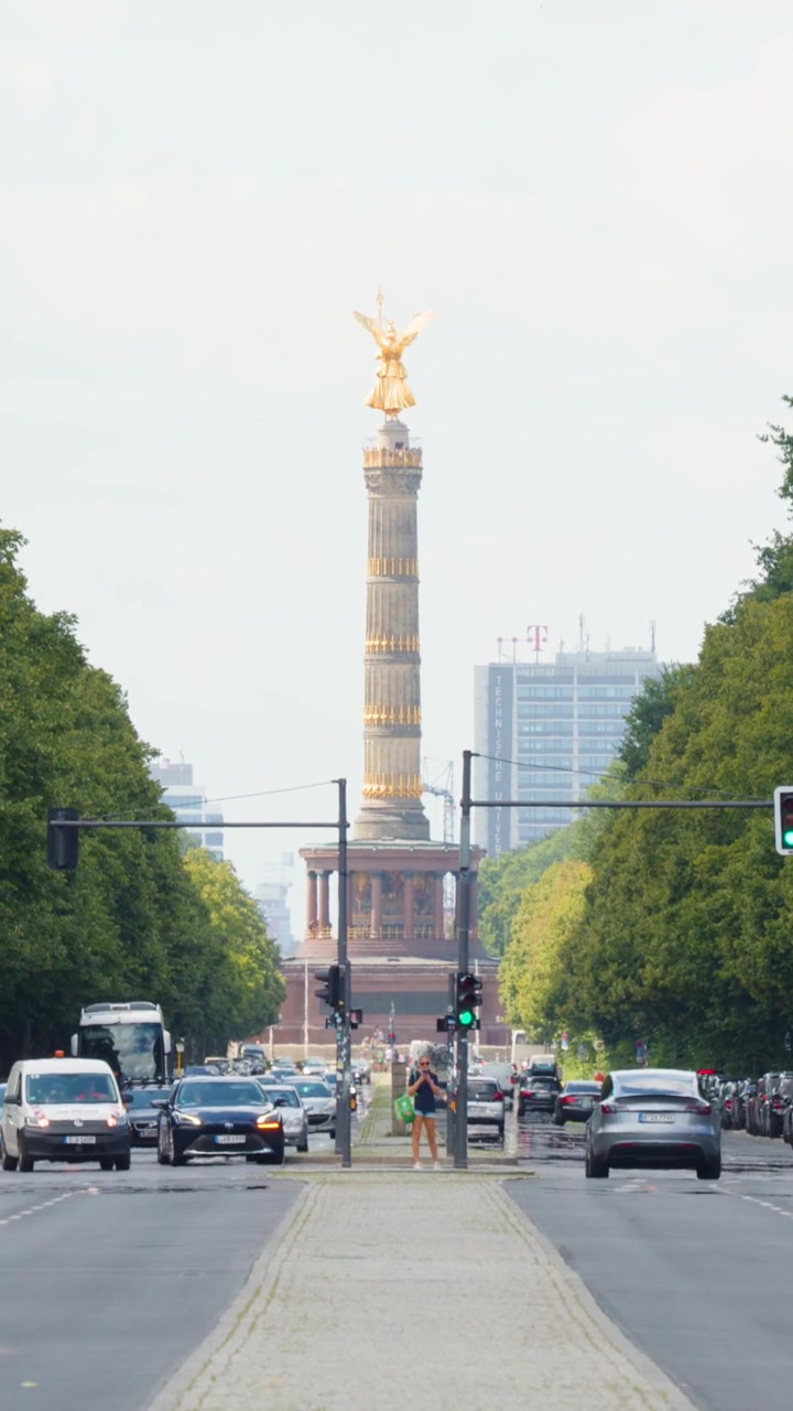 Statisk vidvinkelbillede af Sejrssøjlen i Berlin med biler, der kører langs en træallé.