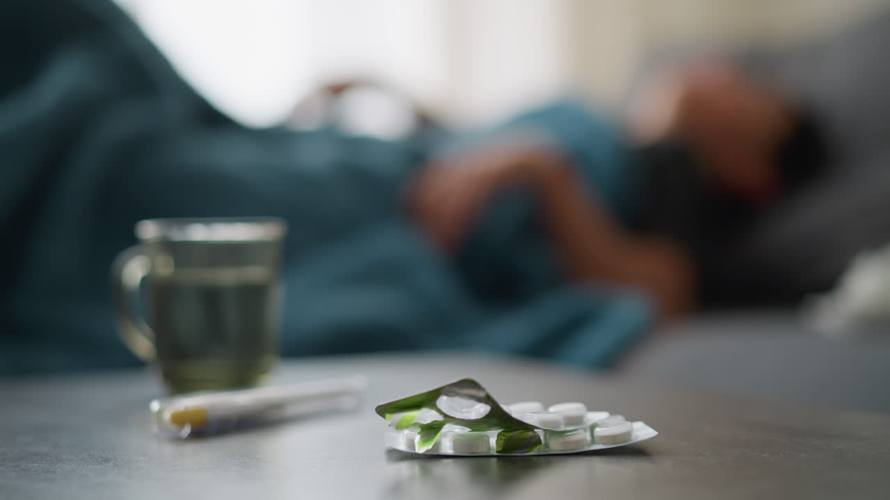 Close-up of medication and glass cup on table with blurred background showing restless person laying down on couch, indicating a sick day with discomfort, recovery, and health focus