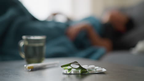 close-up of medication and glass cup on table with blurred background showing restless person laying down on couch, indicating a sick day with discomfort, recovery, and health focus