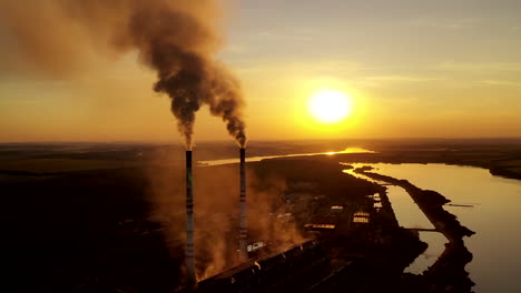 Metallurgy plant in nature at orange sunset. Dark smoke of industrial factory pollute the atmosphere. Evening background of a factory near the river.