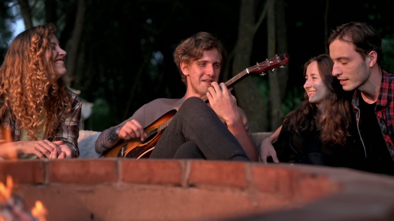 A group of happy young friends near a campfire at glamping, night. Two men and women. Playing guitar. Slow motion