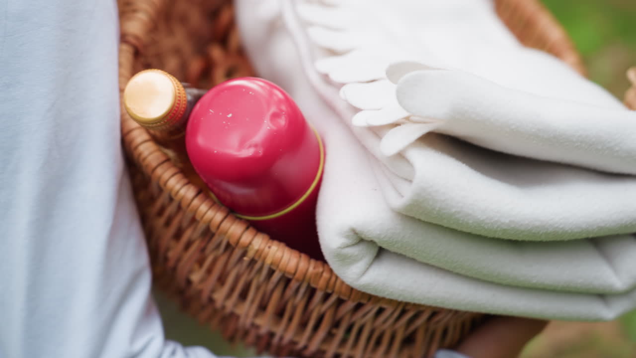 숲길을 따라 걸으며 양손으로 피크닉 바구니를 들고, 접힌 담요와 병을 보여주는 십대 소녀의 하이 앵글 클로즈 뷰로, 기쁨을 상징하는 평화로운 여름 여가 시간을 나타내요.