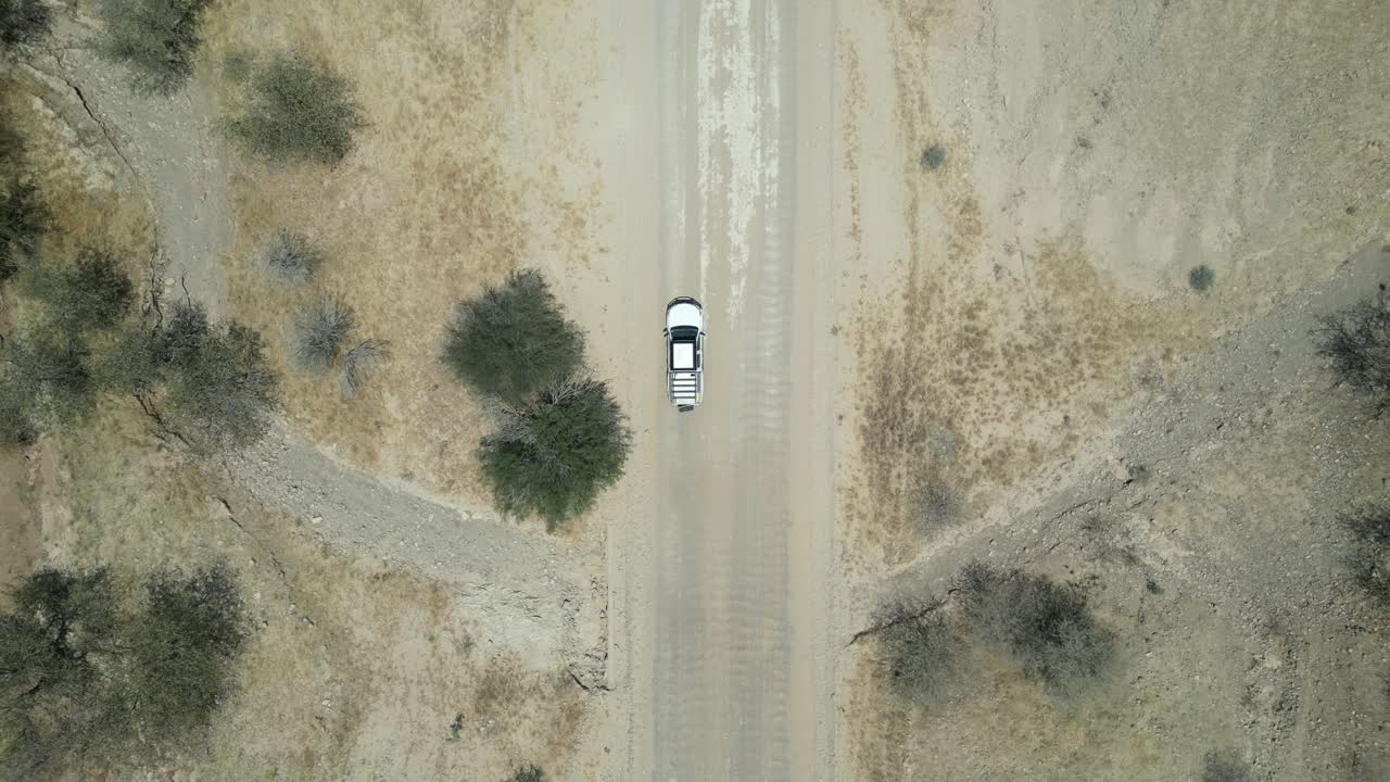 Vista aerea da drone nel deserto della Namibia, con un'auto SUV che percorre una strada stretta, sabbia e alberi verdi ai lati e montagne all'orizzonte in una giornata di sole