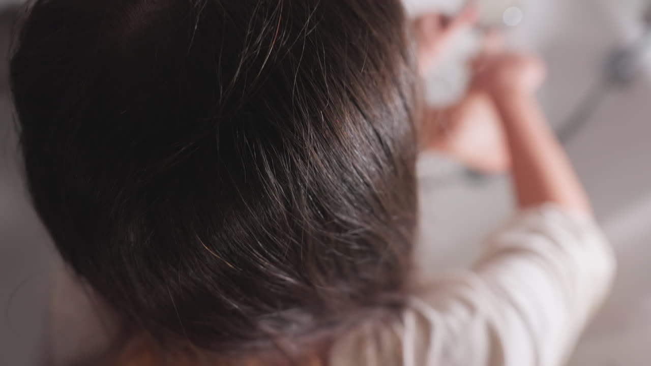 High angle back view shows schoolgirl in bathtub washing feet under running water with hand shower, rinsing toes and hands, long hair visible, clean hygiene routine in tiled bathroom