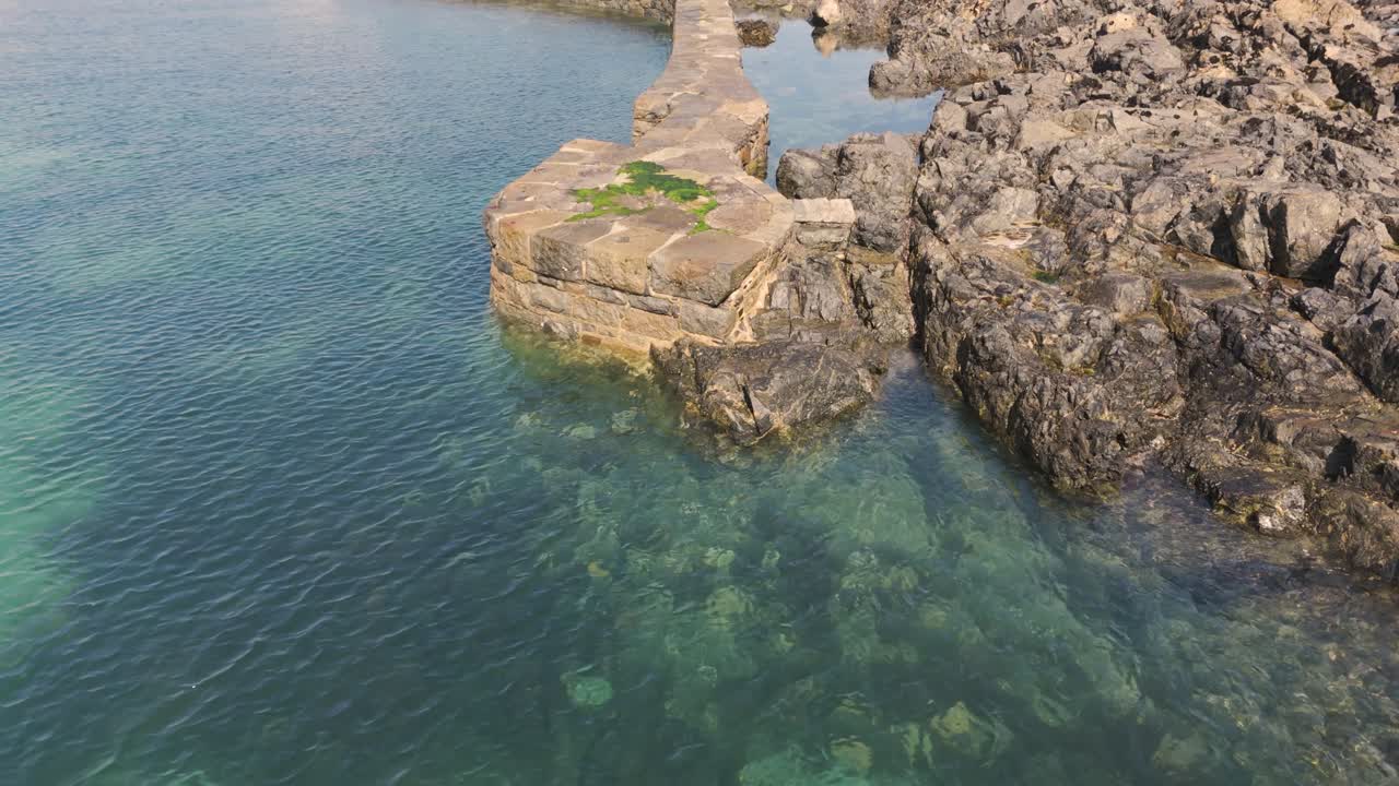 Vecchio muro frangiflutti circondato da acqua calma e limpida e rocce durante la bassa marea, con alghe marine, in una giornata calma e soleggiata.