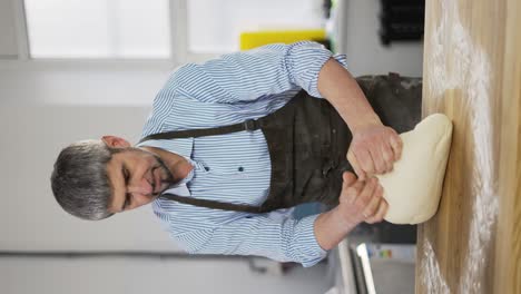 Handsome-male-baker-in-black-apron-kneading-dough,-making-bread-at-the-bakery-kithen.-Vertical-footage