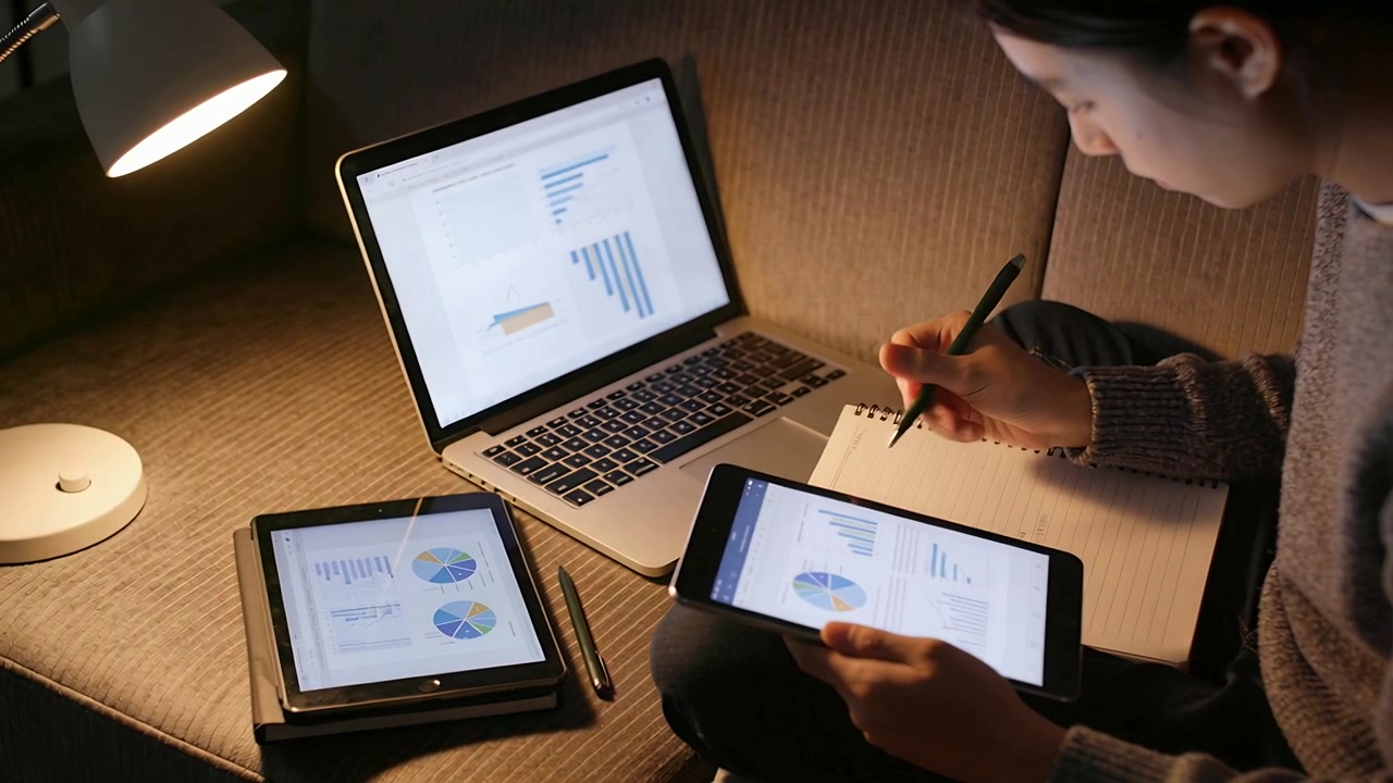 Seeing charts on laptop and tablet, woman in sweater on sofa tapping tablet, writing in notebook