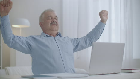 aged man is stretching hands and shoulders during work with laptop sitting at table and typing messages fatigue and overexertion