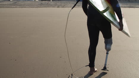 back view of a male surfer with artificial leg walking along beach and holding surfboard under arm