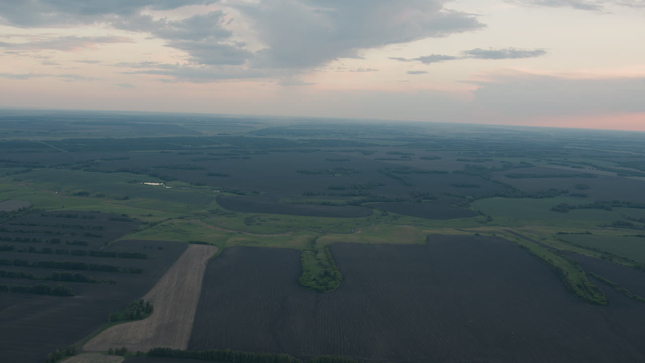 aerial top down perspective of vast patchwork farmland fields with winding waterway corridors and scattered woodland islands under pastel dusk sky capturing timeless serene rural expanse
