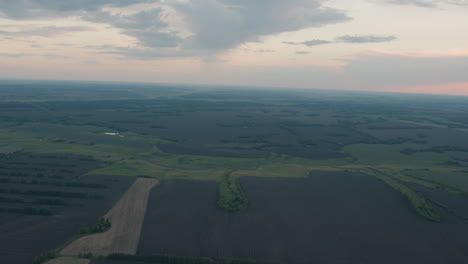 aerial top down perspective of vast patchwork farmland fields with winding waterway corridors and scattered woodland islands under pastel dusk sky capturing timeless serene rural expanse