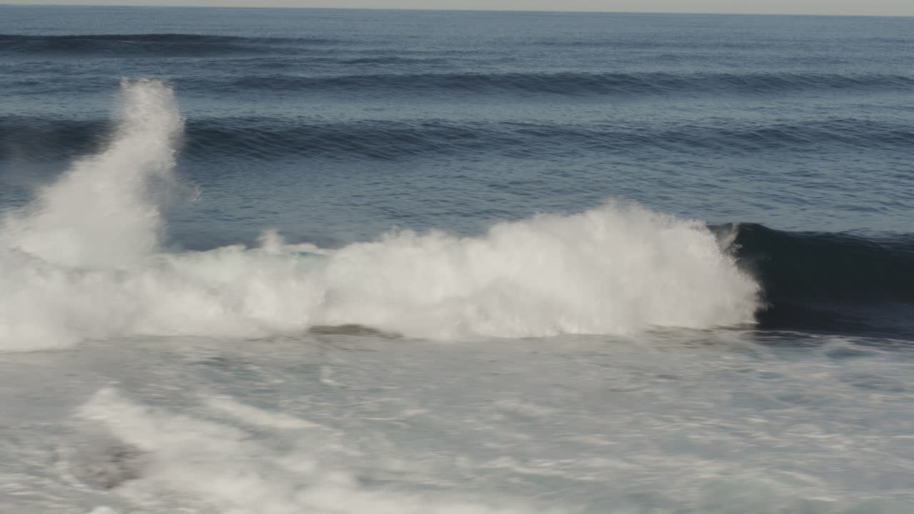 Evening light shines across wave face as surfers float in distant lineup awaiting sets, calm sea scene