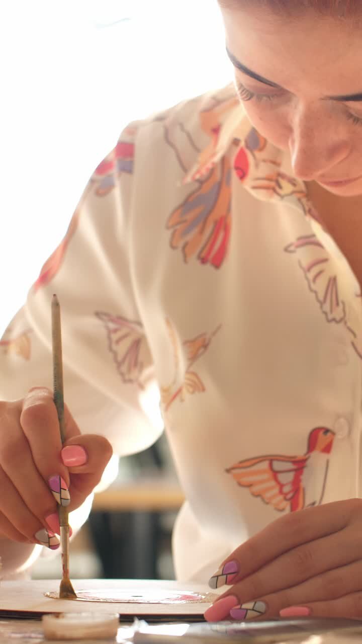 Female artist is focused on painting intricate designs on paper with a brush, showcasing creativity and attention to detail in a bright workspace