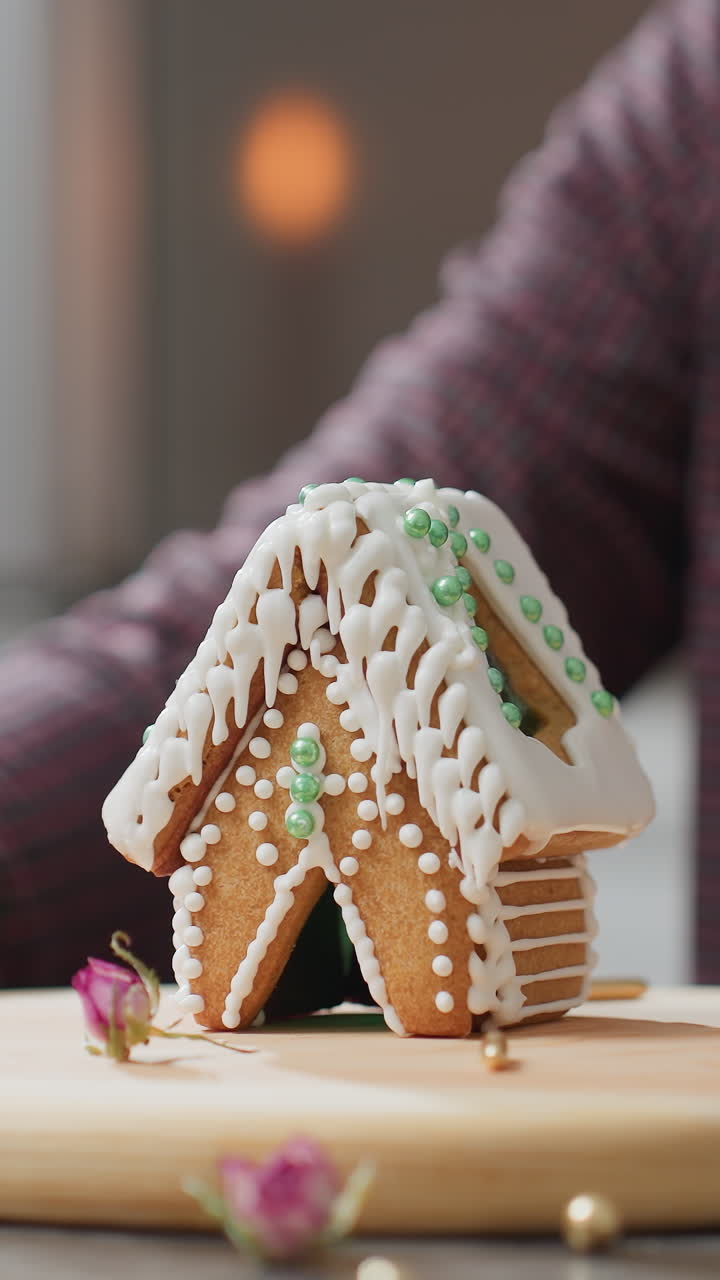 panadero girando cuidadosamente y exhibiendo una casa de pan de jengibre decorada con intrincado hielo blanco y cuentas verdes, bandeja de madera, salpicaduras doradas, flores secas y suave luz de vela en el fondo