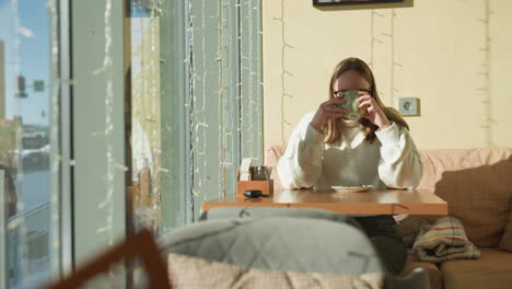 remote worker seated inside cozy cafe sipping morning tea by large window, sunlight casting soft shadows on wall, decorative string lights hanging, reflections of passersby